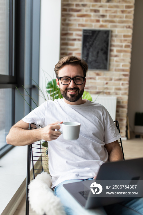 Portrait of a smiling man relaxing on chair near window using laptop and holding coffee cup at home
