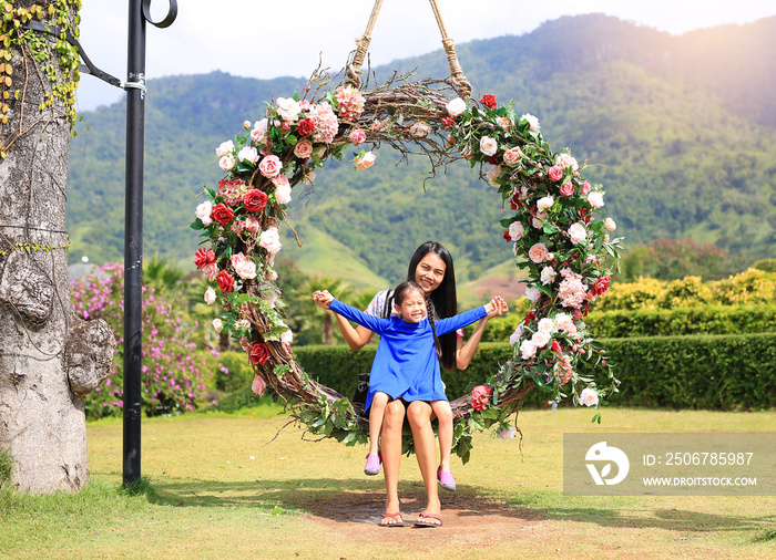 Asian mother and her daughter sitting on beautiful basket swing with the colorful roses flower in th