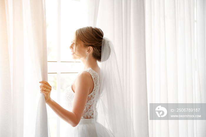 Beautiful young bride near window before wedding ceremony at home