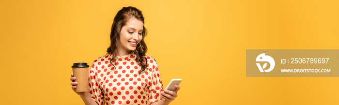 panoramic shot of happy young woman holding coffee to go and using smartphone isolated on yellow