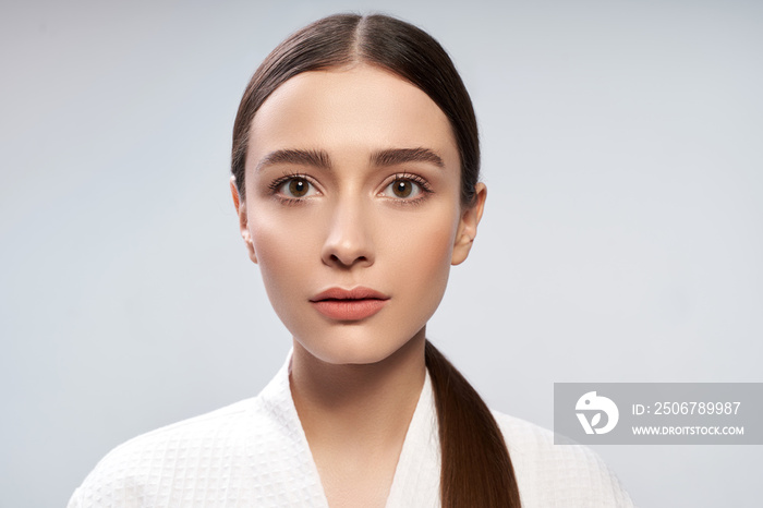 Attractive young woman in bathrobe standing against light blue background