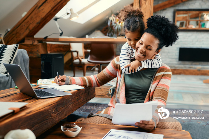 Small black girl embraces her busy mother who is working at home.