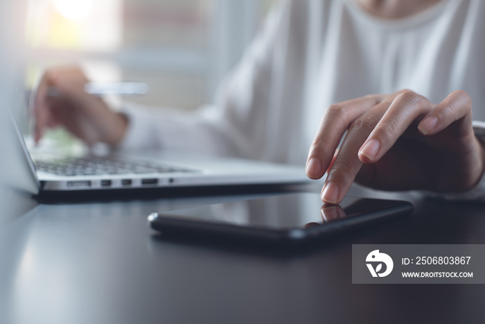 Woman hand touching on mobile phone screen and working on laptop computer on table from home office