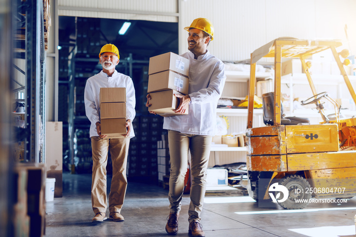 Two blue collar workers in white uniforms and with yellow helmets on heads relocating heavy boxes in