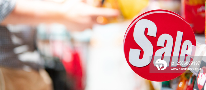 Red sale label on product shelf in supermarket with blurred male shopper choosing food package in th