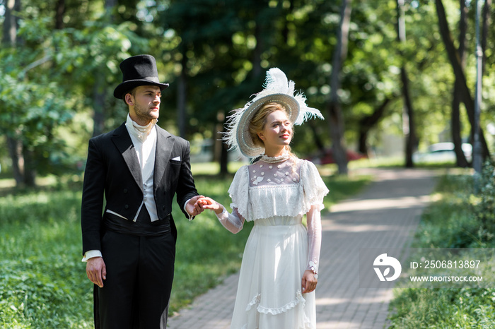 victorian man and woman in hats holding hands while standing outside near green trees