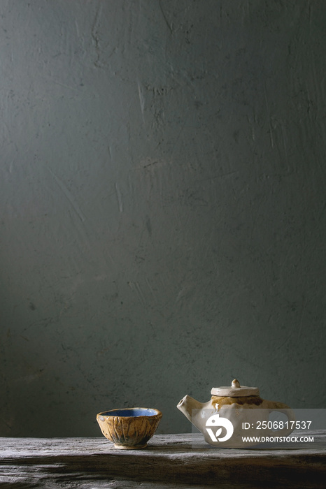 Craft handmade ceramic teapot and cup for tea ceremony standing on old wooden shelf in dark room.