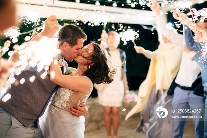 groom and bride kissing among guests with sparklers at the wedding party