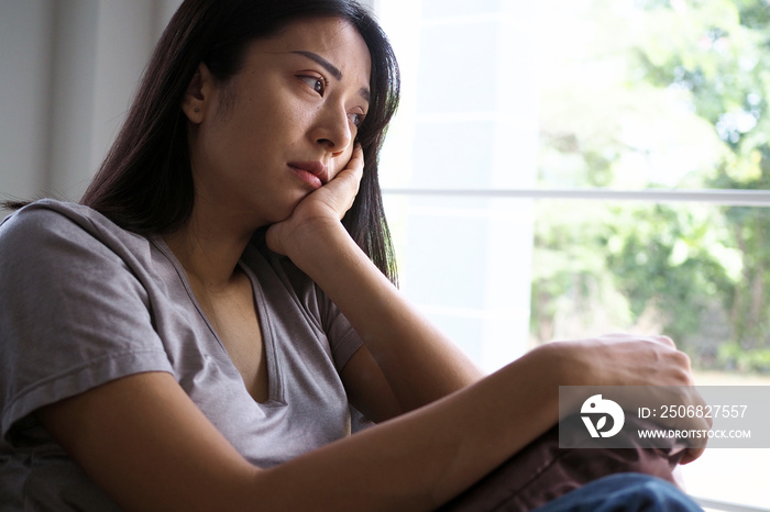 Asian woman sitting inside the house looking out at the window. Woman confused, disappointed, sad an