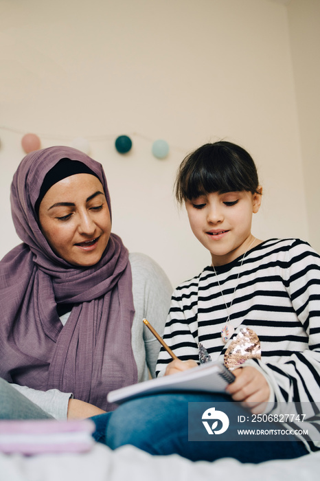 Low angle view of mother assisting daughter in doing homework on bed at home