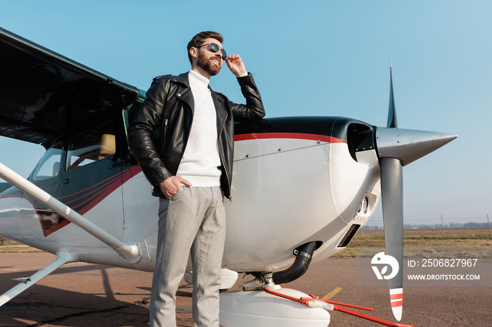 low angle view of happy pilot in leather jacket adjusting sunglasses and standing with hand in pocke