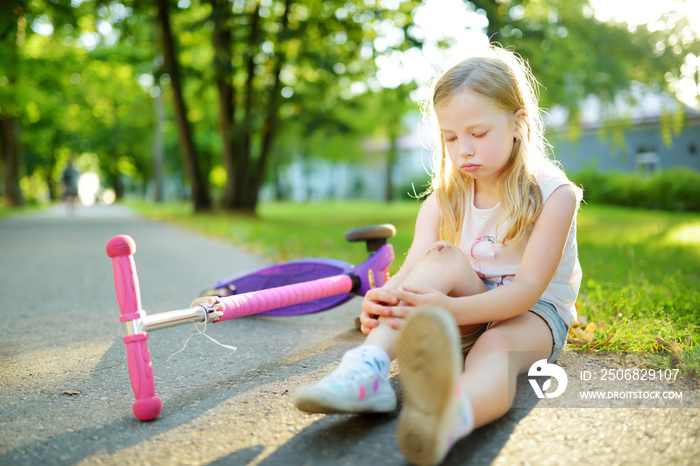 Cute little girl sitting on the ground after falling off her scooter at summer park. Child getting h