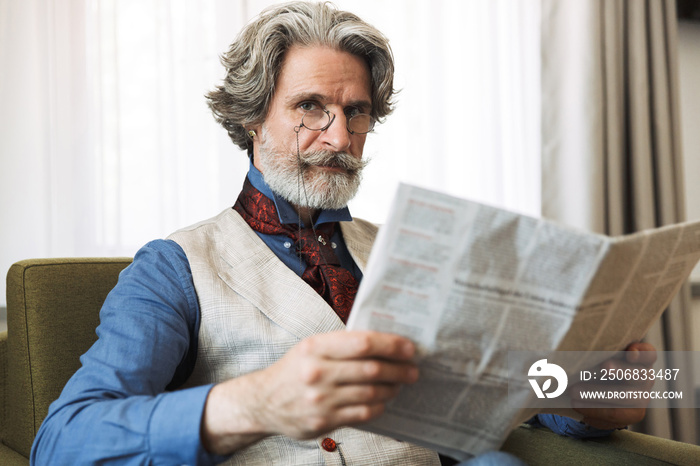 Portrait of bearded adult businessman reading newspaper while sitting on armchair in hotel apartment