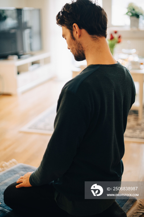 Mature man praying while sitting on carpet in living room