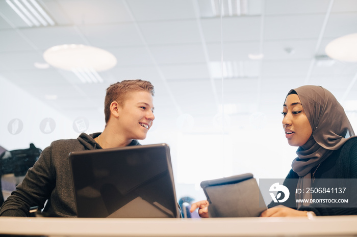 Smiling classmates using technologies while sitting in classroom at university