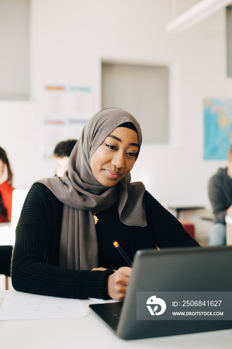 Smiling teenage student in hijab using laptop at classroom