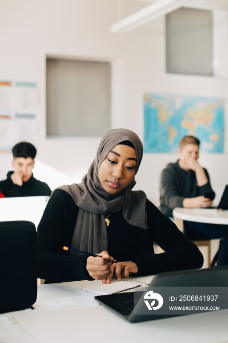 Teenage girl wearing hijab is writing in book while using laptop at desk in classroom