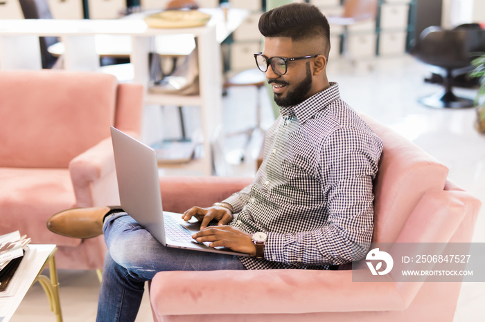 Casual young indian man using laptop with happy on sofa in modern office