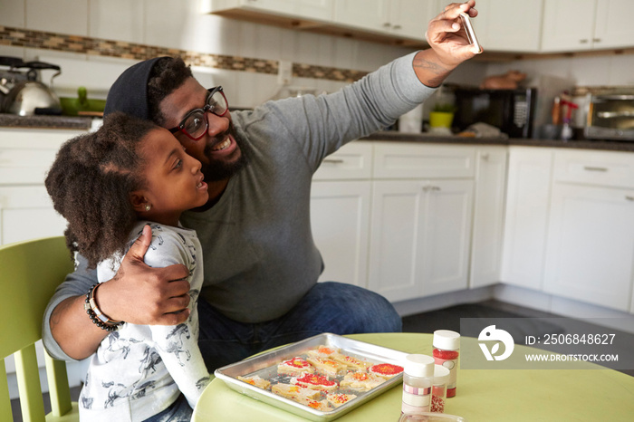 Father and daughter in kitchen, baking, father taking selfie of them both using smartphone