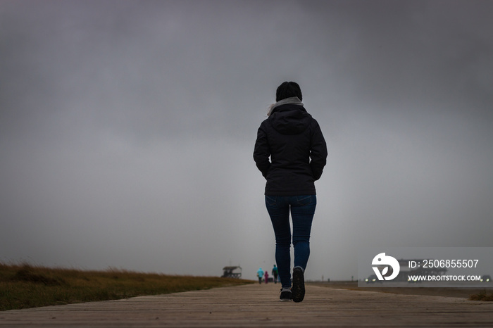 Unrecognizable woman walking away on a wooden footpath at north sea beach.