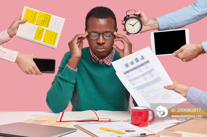 Concentrated black man keeps hands on temples, wears spectacles, green jumper, surrounded with paper