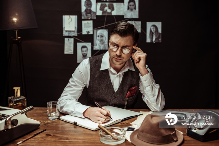 Pensive detective in glasses sitting at table and writing in notebook