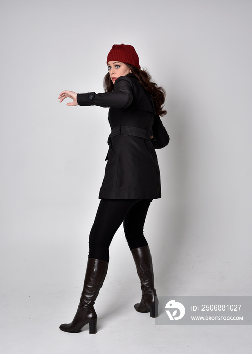 full length portrait of a pretty brunette girl wearing a black coat, leather boots and red beanie. S