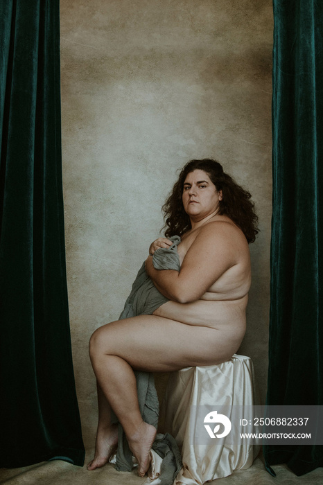 portrait of a plus size woman with curly hair sitting on a stool looking at camera clutching gray fa