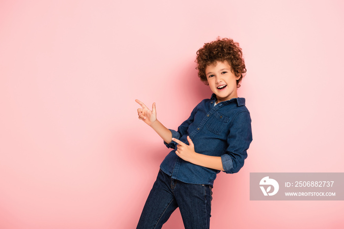 excited curly boy in denim shirt pointing with fingers on pink