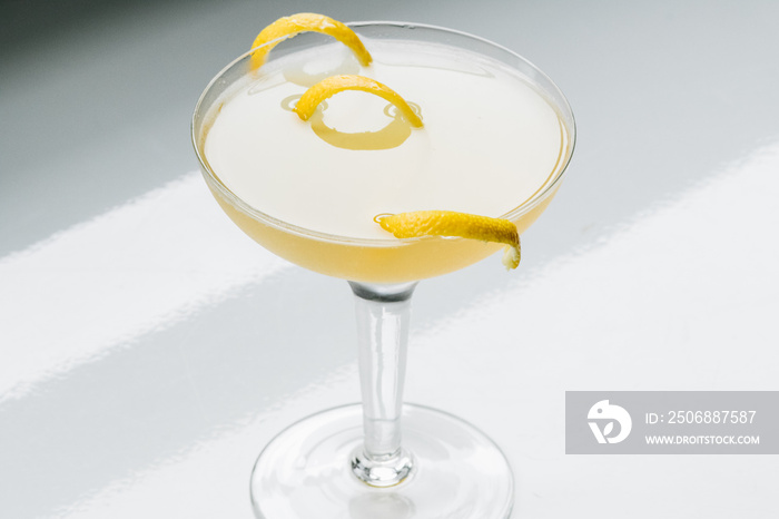 yellow cocktail in coupe glass with long curly lemon twist against white background