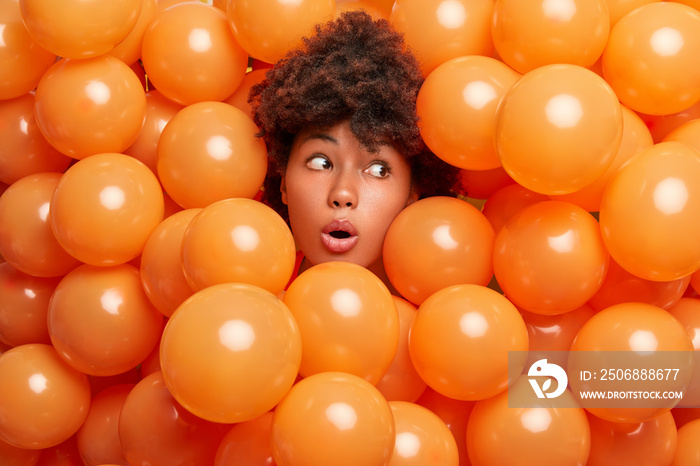 Shocked excited Afro American girl with curly hair surrounded by holiday inflated balloons looks awa