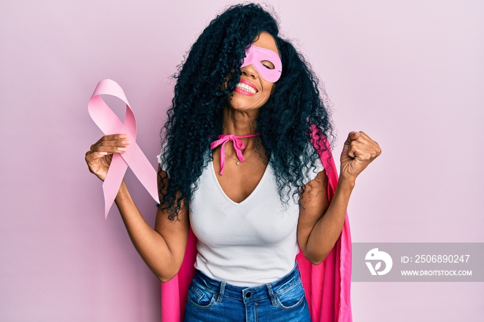 Middle age african american woman wearing super hero costume holding pink cancer ribbon screaming pr