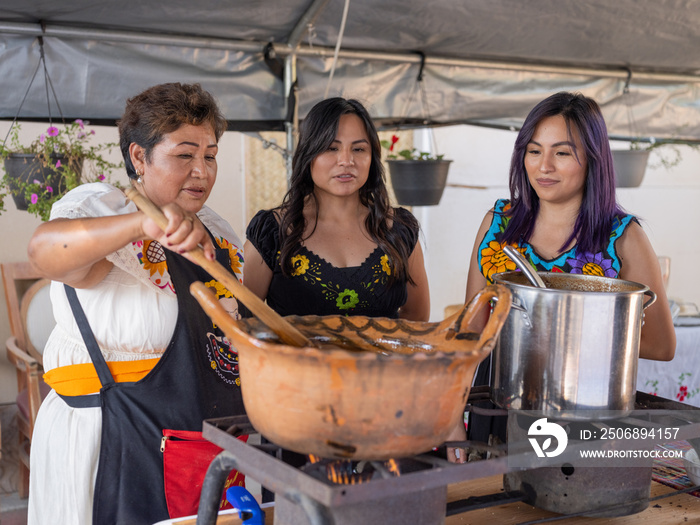 Indigenous daughters speak to mom as she stirs food in a clay pot