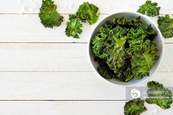 Healthy kale chips in a bowl. Above scene, corner border on a white wood background. Copy space.