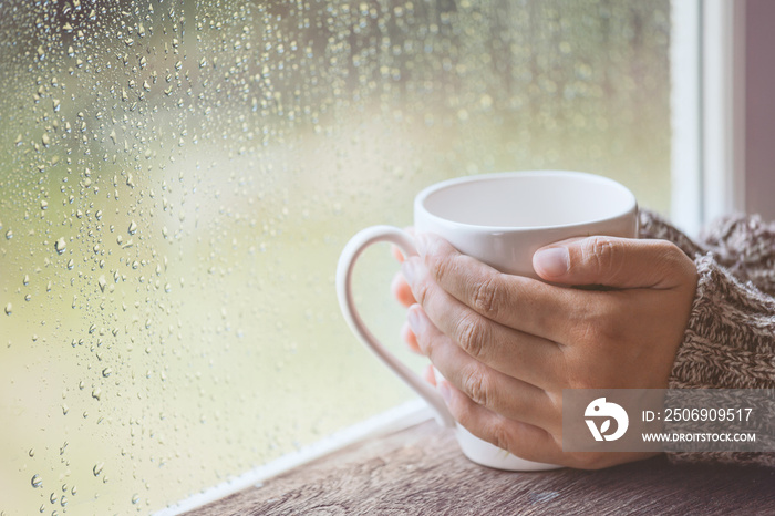 在雨天的窗户背景上，复古色调的女人手里拿着一杯咖啡或茶
