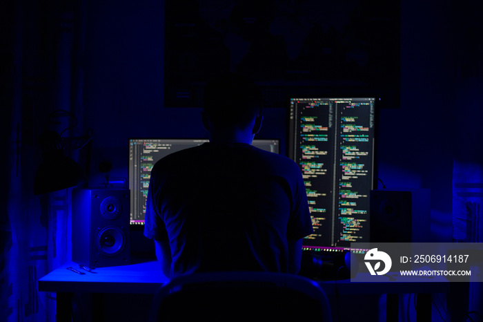 A man sits at a computer in a room at a table at night with blue lighting and programs