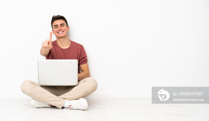 Teenager man sitting on the flor with his laptop showing and lifting a finger