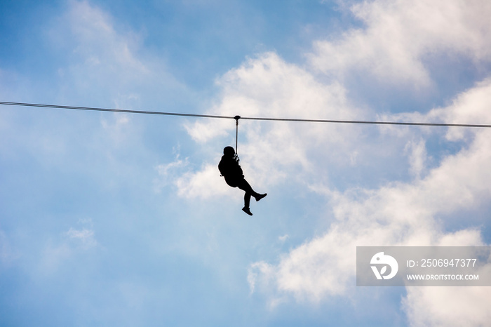 Silhouette of a hanging man on a zip line in front of a cloudy blue sky background