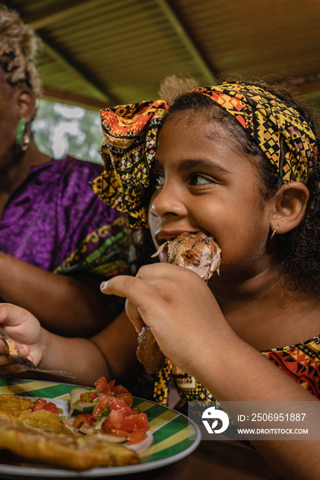 Imagen vertical de una niña afrocaribeña disfrutando de un delicioso trozo de pollo caribeño sentada