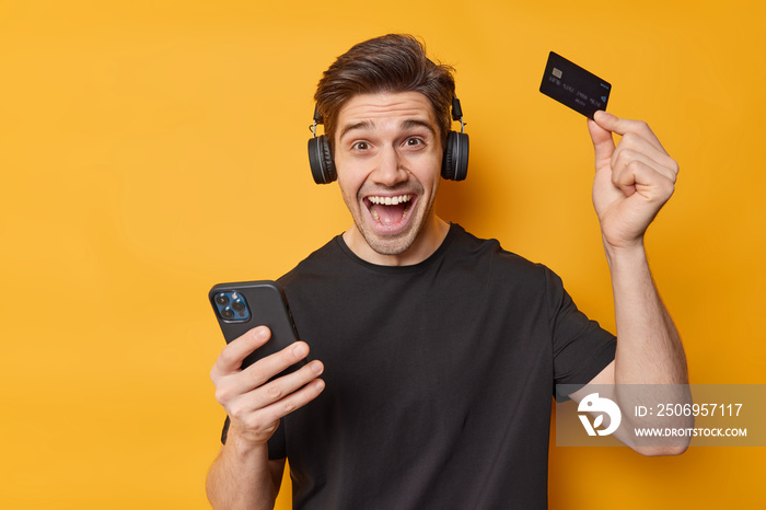 Joyful adult man holds mobile phone credit card recommends bank or shopping application listens musi