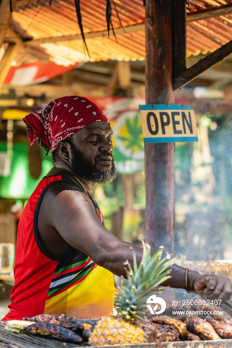 Imagen vertical de un hombre afro caribeño con un pañuelo en su cabeza cocinando pollo y otras fruta