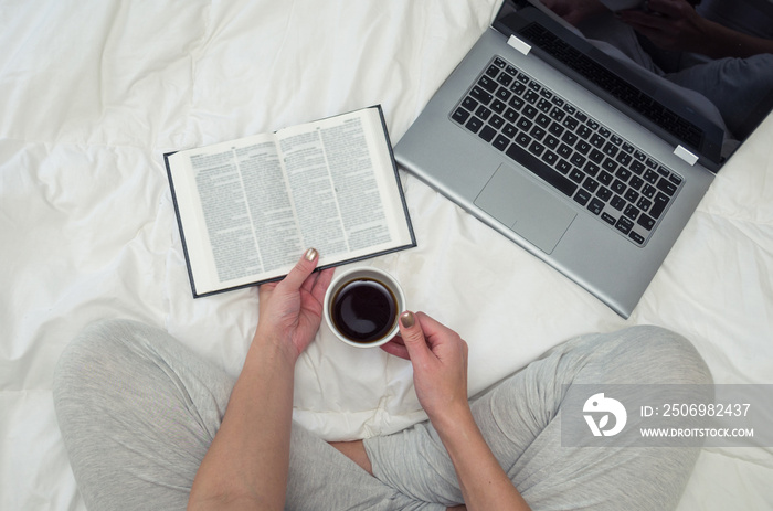 Mão de mulher segurando xícara de café, livro e notebook na cama, visto de cima.