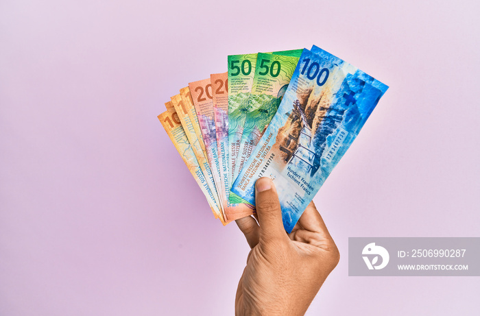 Hispanic hand holding swiss franc banknotes over isolated pink background.