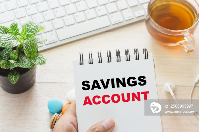 Man holding SAVINGS ACCOUNT message on book and keyboard with a hot cup of tea, macaroon on the tabl