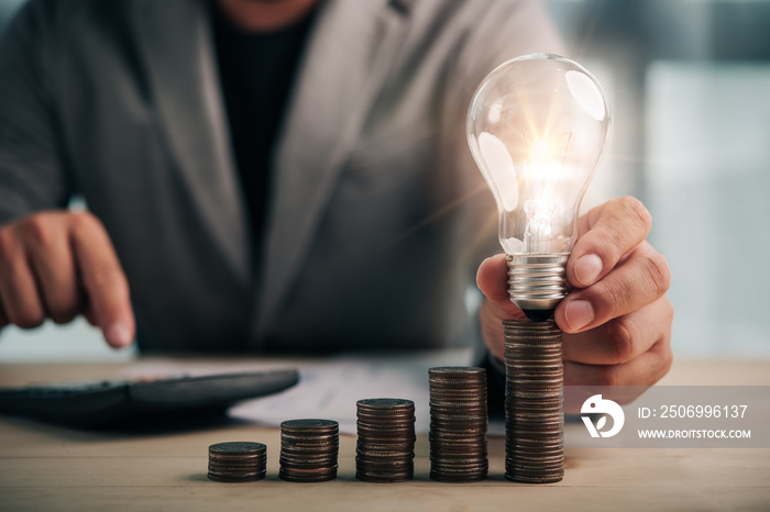 businessman hand holding lightbulb with coins stacking on desk.saving energy and money concept.