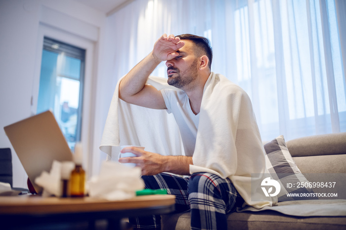 Hardworking employee in pajamas and covered with blanket having fever. Man holding mug with tea and 