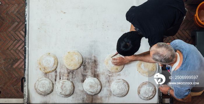 Artisan baker teaches his apprentice to slice yeast bread dough before bake in electric oven.