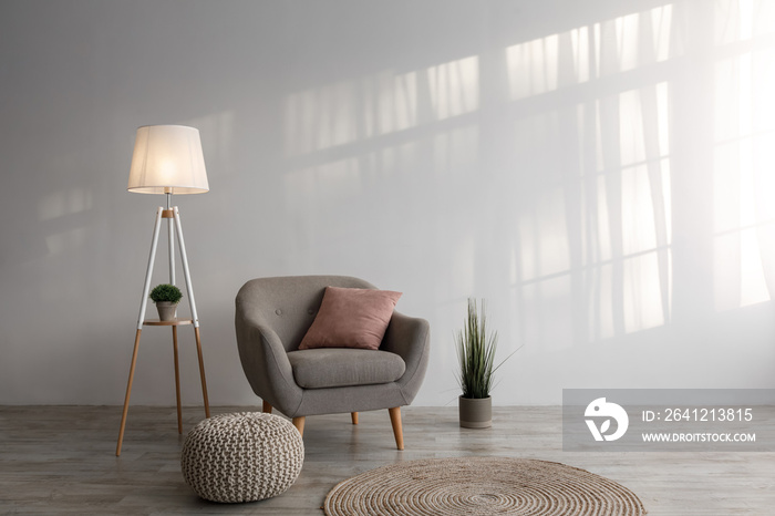 Armchair with pillow, lamp, plant in pot, ottoman and round carpet on floor on gray wall background 