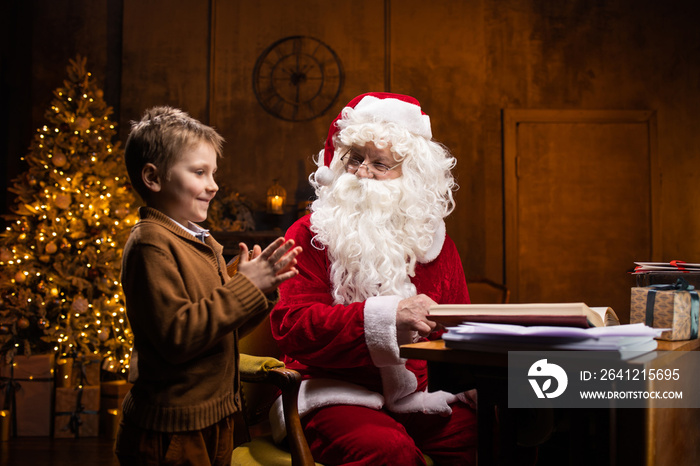 Santa Claus and little boy. Cheerful Santa is working while sitting at the table. Fireplace and Chri