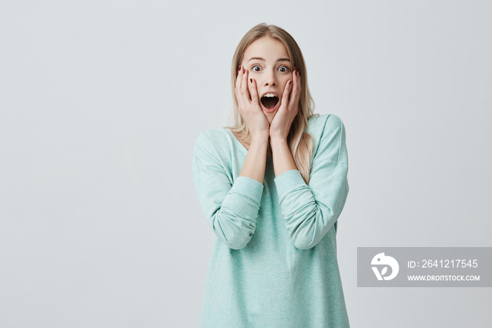 Surprised pretty young female with long dyed blonde hair, looks with opened mouth at camera, shocked